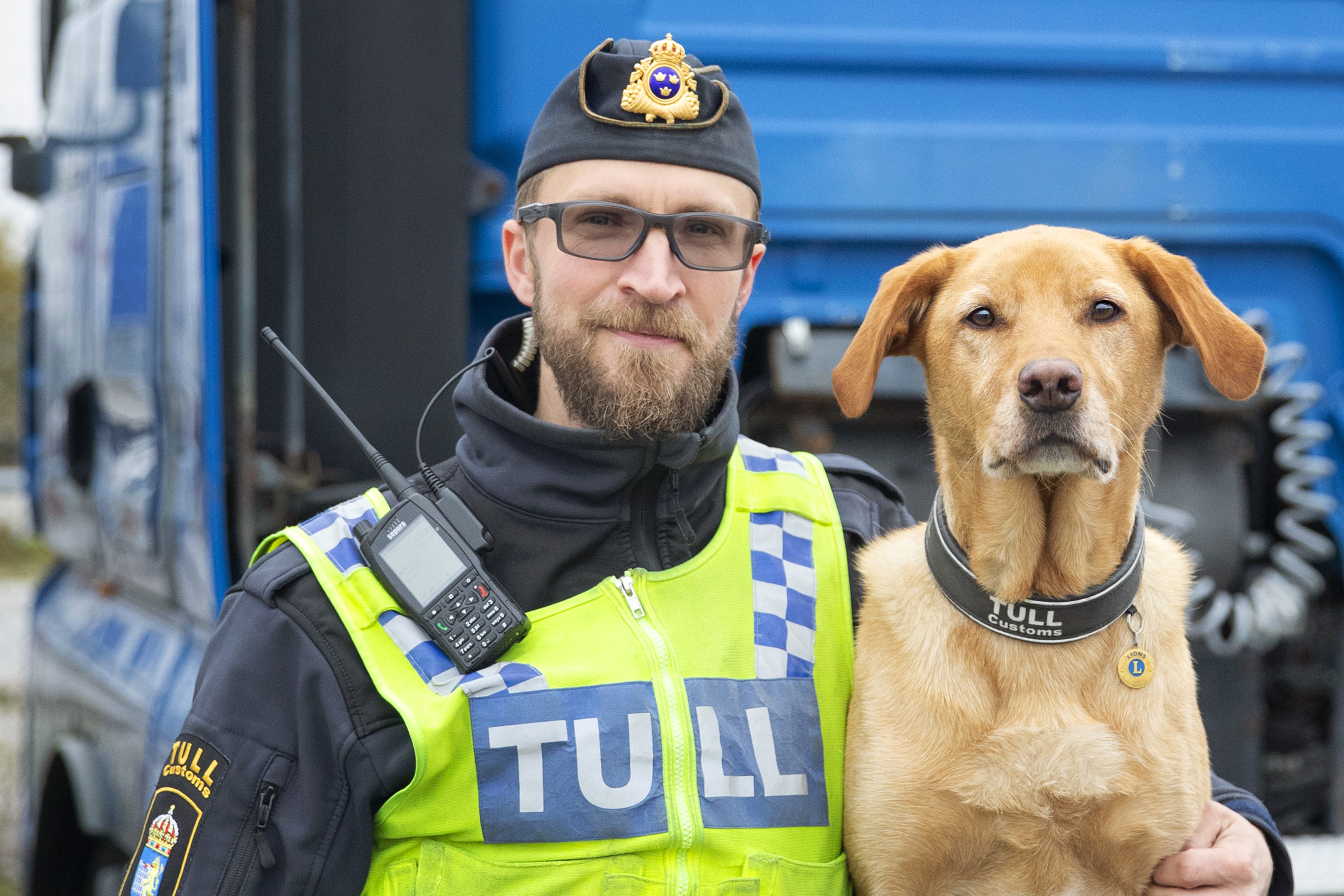 Tullinspektör i uniform håller om en labrador retriever som har ett halsband det står "Tull Customs" på. Fotograf Mette Ottosson.