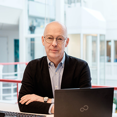 En bild på Dan som sitter vid en laptop i en ljus och luftig inomhusmiljö och tittar in i kameran med en mjuk blick. Det är en äldre man utan hår, med runda glasögon, blå skjorta och en svart kofta.
