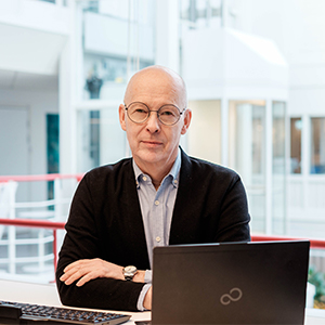 En bild på Dan som sitter vid en laptop i en ljus och luftig inomhusmiljö och tittar in i kameran med en mjuk blick. Det är en äldre man utan hår, med runda glasögon, blå skjorta och en svart kofta.