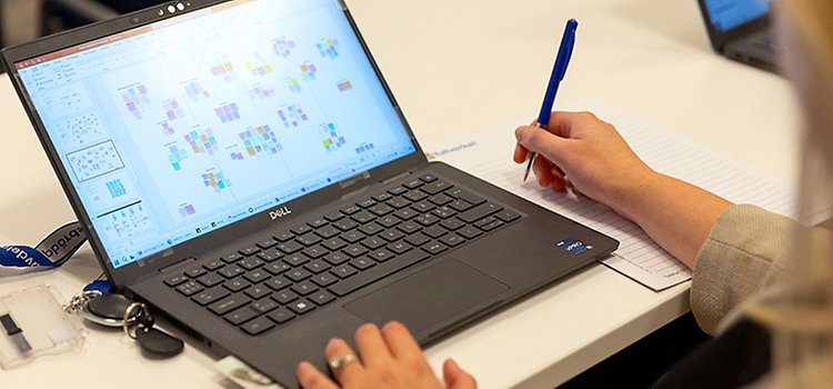 En person jobbar på en bärbar dator, skriver med en blå penna på ett papper intill.